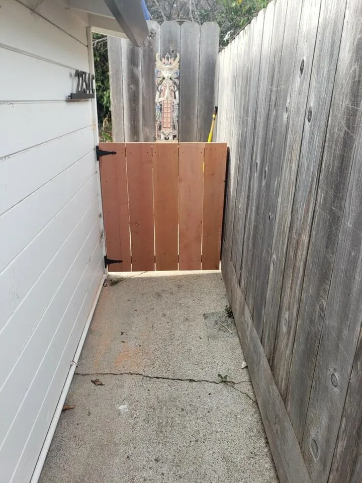 Closed vertical-board wooden gate across a narrow passage between house siding and a timber fence.