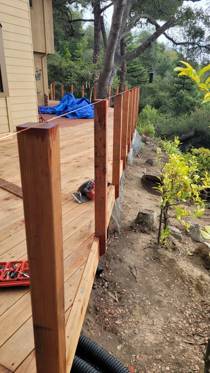 A row of wood railing posts being aligned along the unfinished deck edge beside a wooded slope.