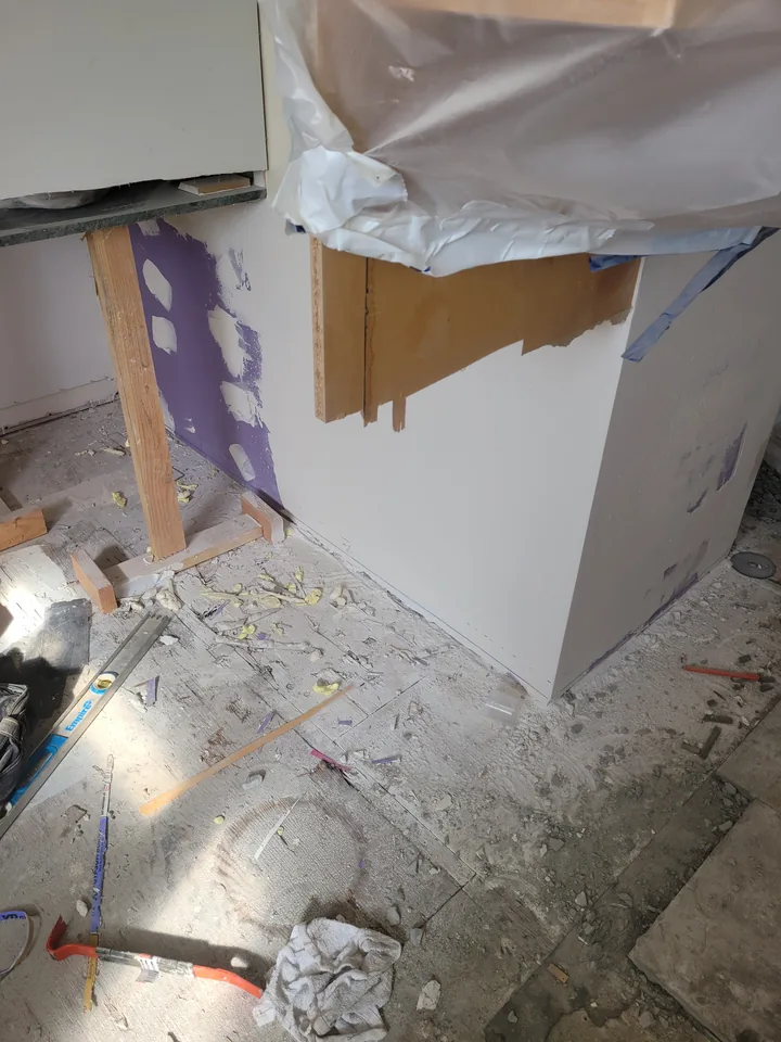 Kitchen wall and cabinet area with drywall patches, protective plastic, tools, and unfinished flooring.